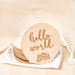 Small round wooden disc with the words 'hello world' and a graphic of a rainbow etched into the wood.
